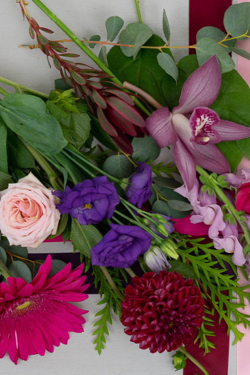 A vibrant bouquet of flowers featuring shades of pink, fuchsia, burgundy, and purple, with a variety of foliages.