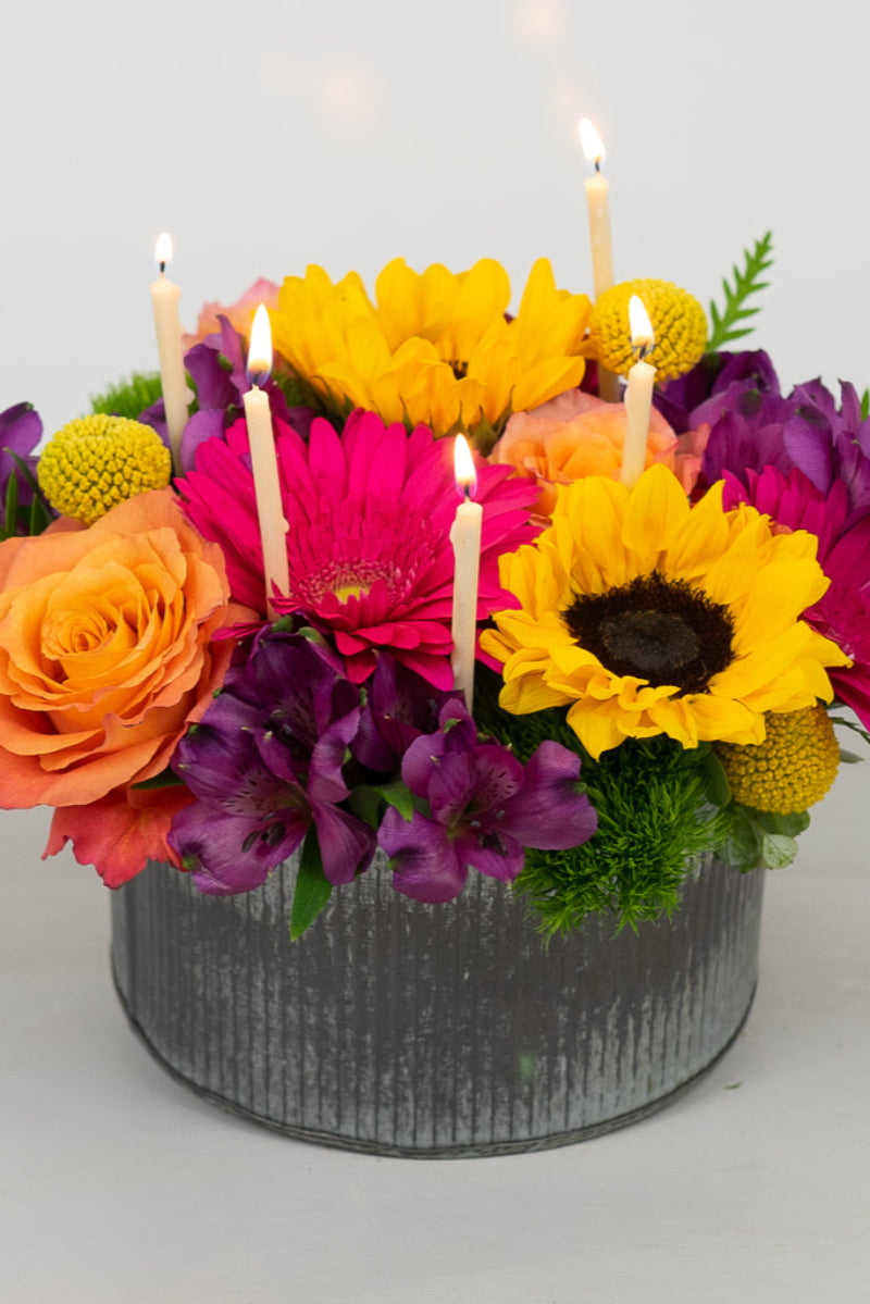 A colorful floral arrangement featuring roses, gerbera daisies, and sunflowers with candles, presented in a tin container.