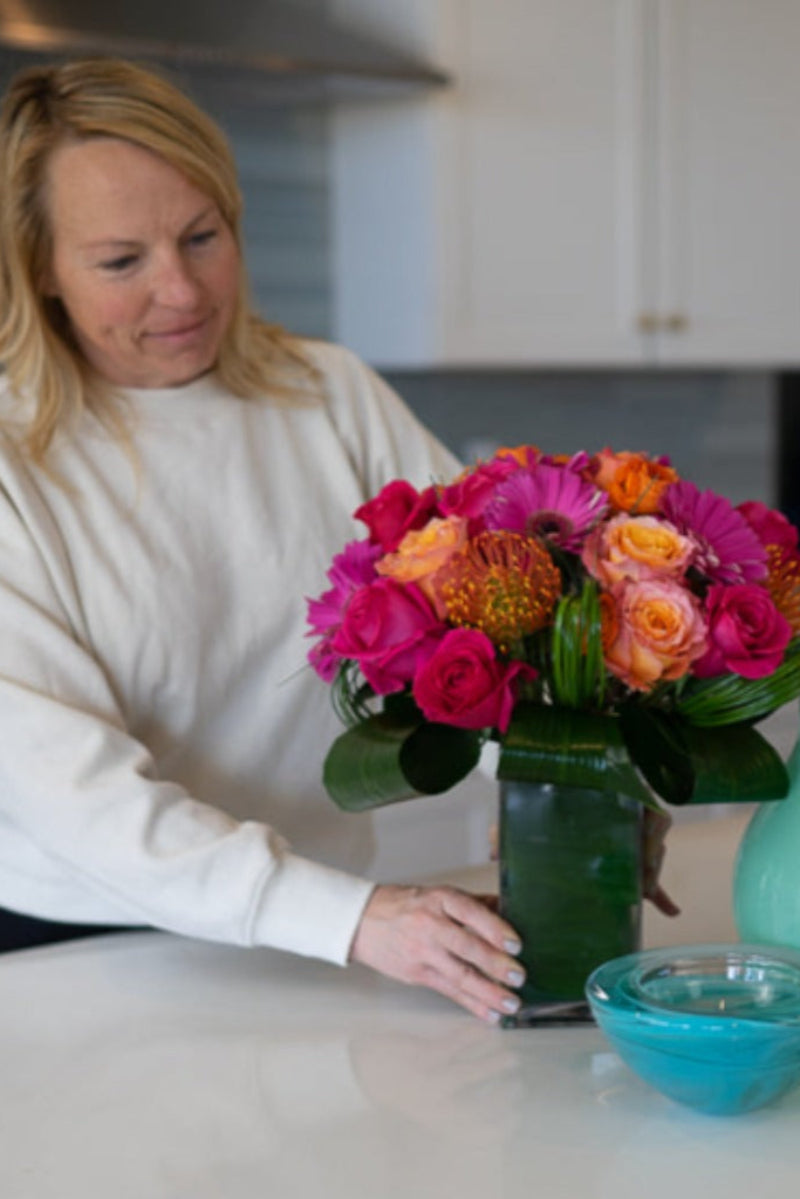 A person arranging a colorful bouquet of flowers in a vase on a kitchen counter.
