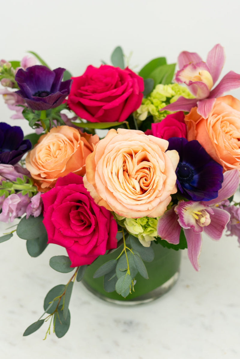 A vibrant floral arrangement featuring roses, snapdragons, anemone, and orchids in a cylinder vase with a mix of pink, purple, and ruby tones.