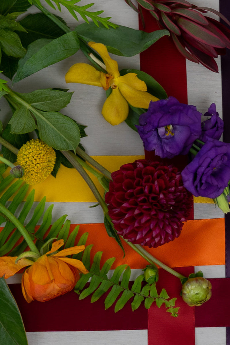 A vibrant bouquet featuring a mix of purple flowers, golden yellow leaves, orange flowers, and deep green foliage, arranged against a white and purple striped background.