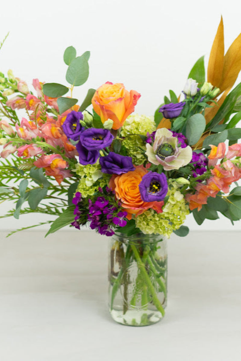 A colorful bouquet of flowers arranged in a clear glass mason jar vase, featuring shades of purple, orange, green, and yellow.