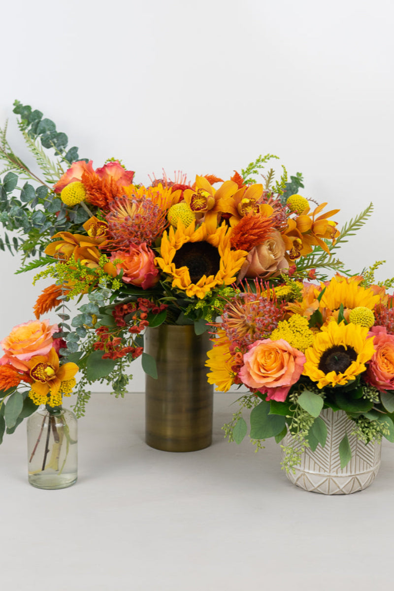 A collection of four floral arrangements with a mix of sunflowers, roses, and other foliage in shades of orange, yellow, and green, arranged in different sized vases against a white background.