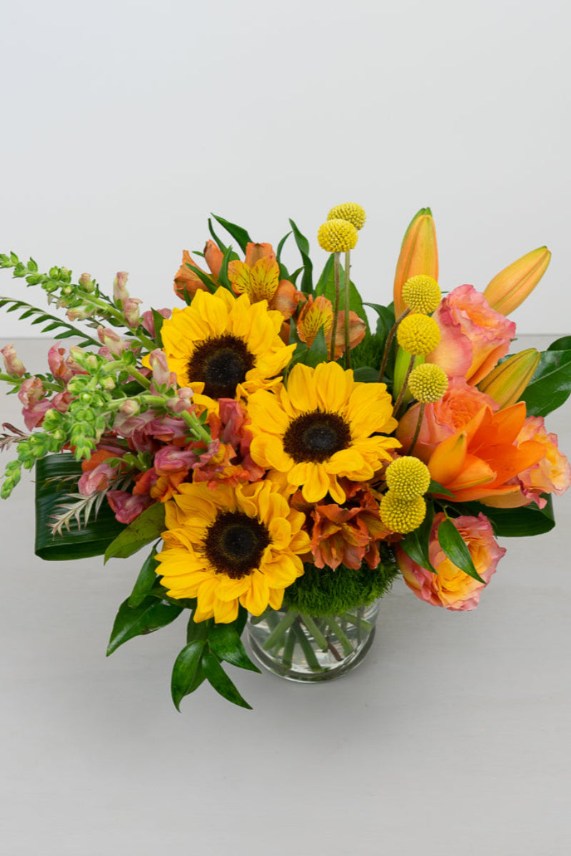 A vibrant bouquet of sunflowers, snapdragons, roses, and lilies arranged in a clear vase, with a mix of yellow, orange, and green colors.
