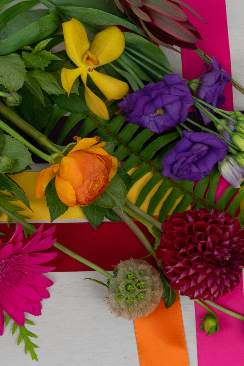 A colorful bouquet of various flowers including yellows, purples, reds, and oranges, alongside green foliage and a red berry, arranged against a checkered background.