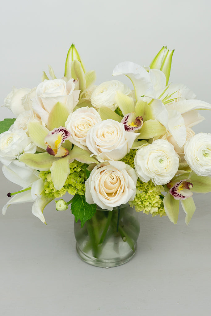 A bouquet of white roses, orchids, ranunculus, and lilies arranged in a clear glass vase, with green premium foliage.