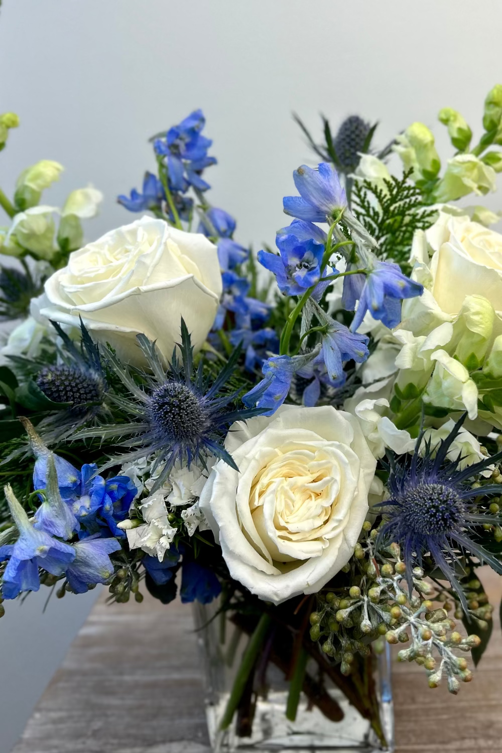 Bouquet of white roses, blue flowers, and thistles in a clear vase against a light gray background.