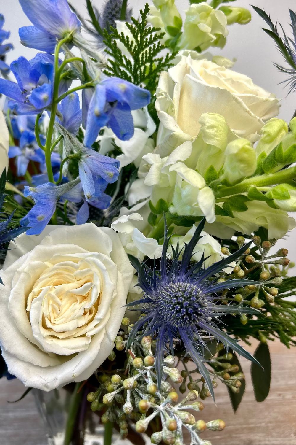 Bouquet of flowers with white roses, blue delphinium, and greenery on a wooden surface.