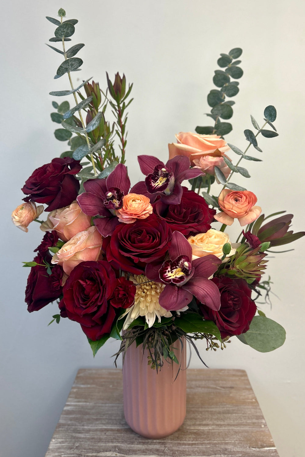 Luxe Blush &amp; Bordeaux Bloom Vase featuring burgundy orchids, additional roses, café latte cremones, peach ranunculus, and burgundy foliage arranged in a taller blush vase ready for delivery