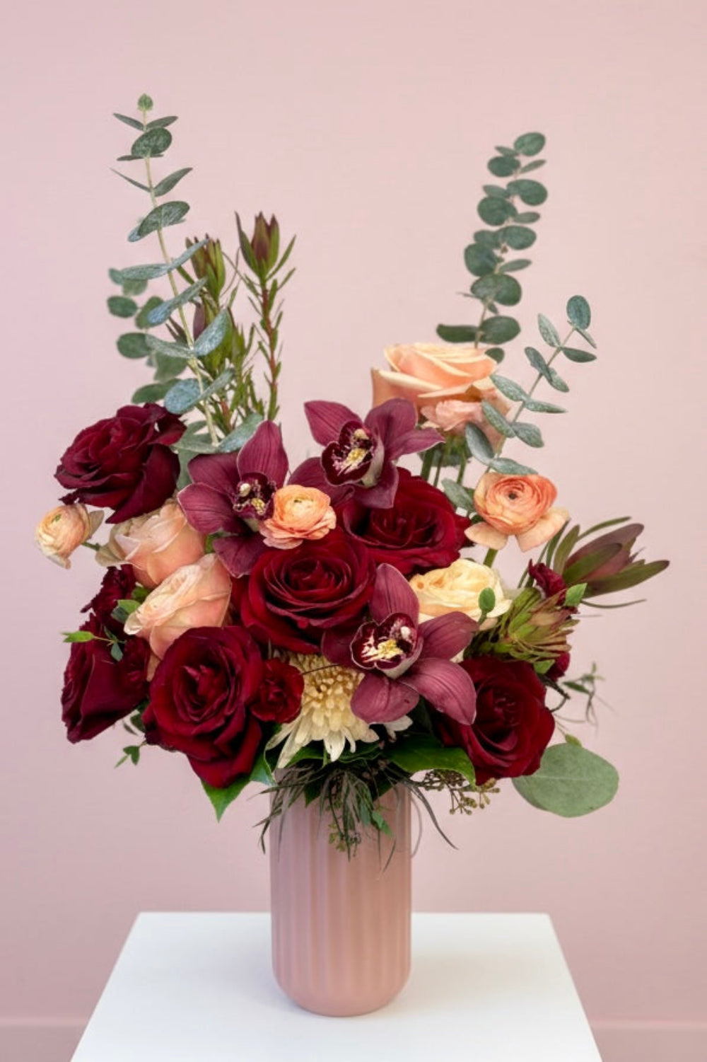 Luxe Blush & Bordeaux Bloom Vase featuring burgundy orchids, additional roses, café latte cremones, peach ranunculus, and burgundy foliage arranged in a taller blush vase.