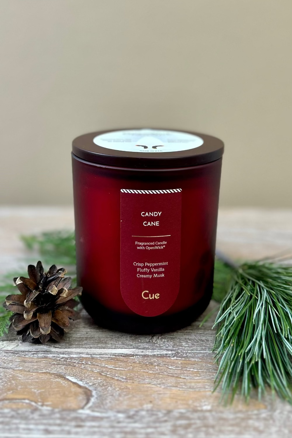 Red candle labeled &#39;Candy Cane&#39; by Cue on a wooden surface with greenery and a pine cone.