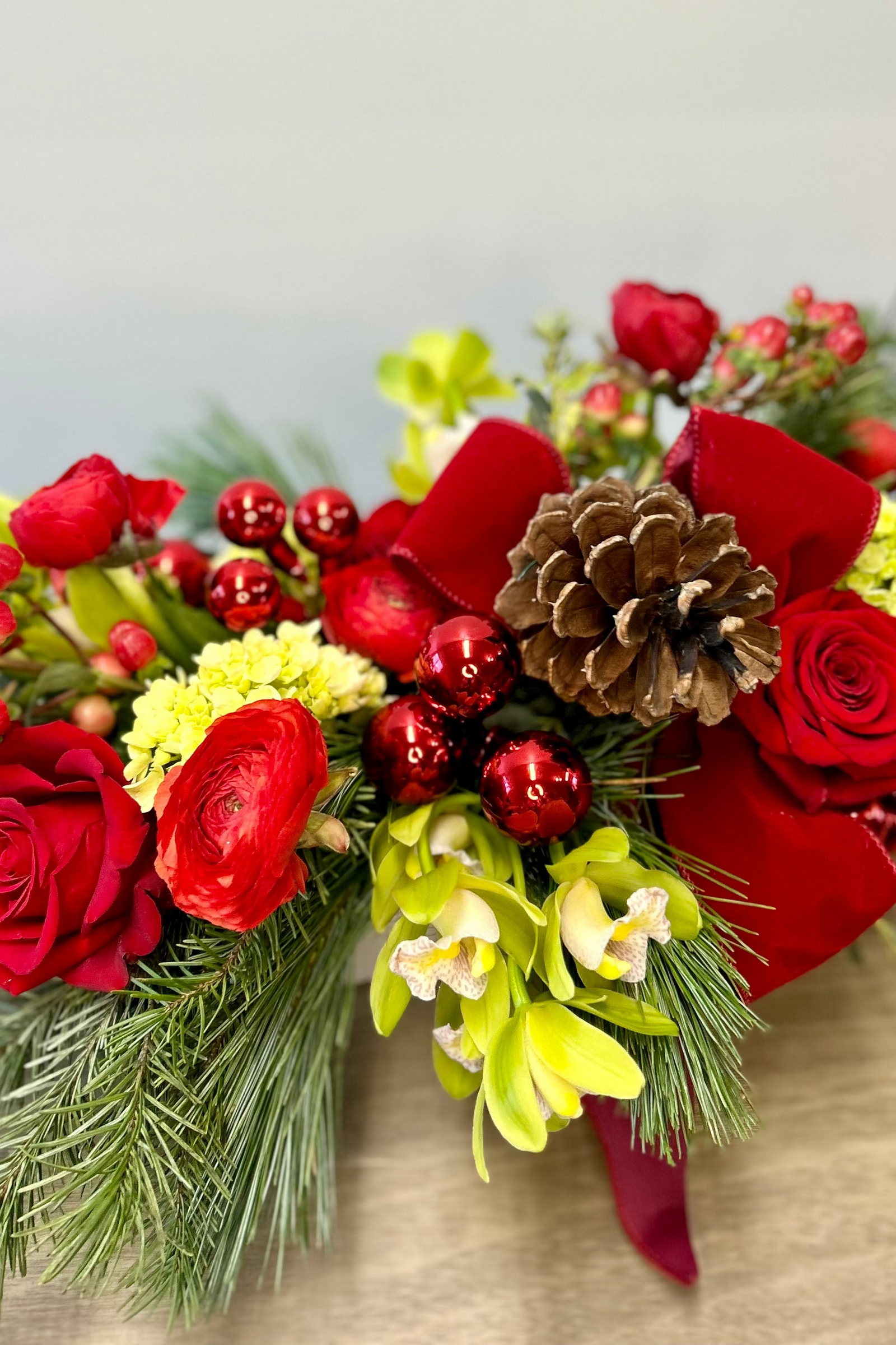 A festive Christmas centerpiece featuring a variety of decorative flowers including red roses, green hydrangea, and white orchids, arranged with evergreens and decorative pine cones.