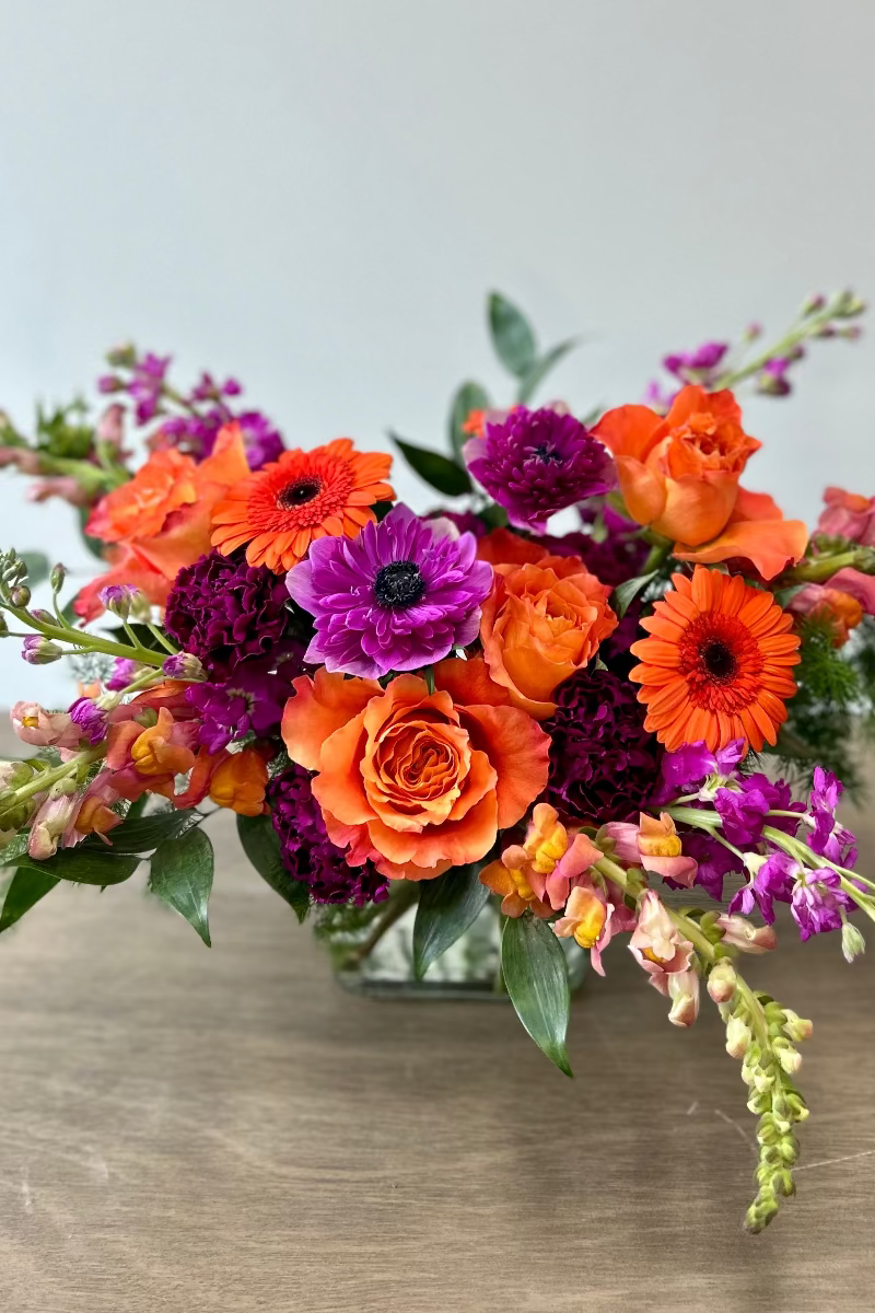 Colorful flower arrangement with orange, purple, and pink flowers in a clear vase on a wooden surface.