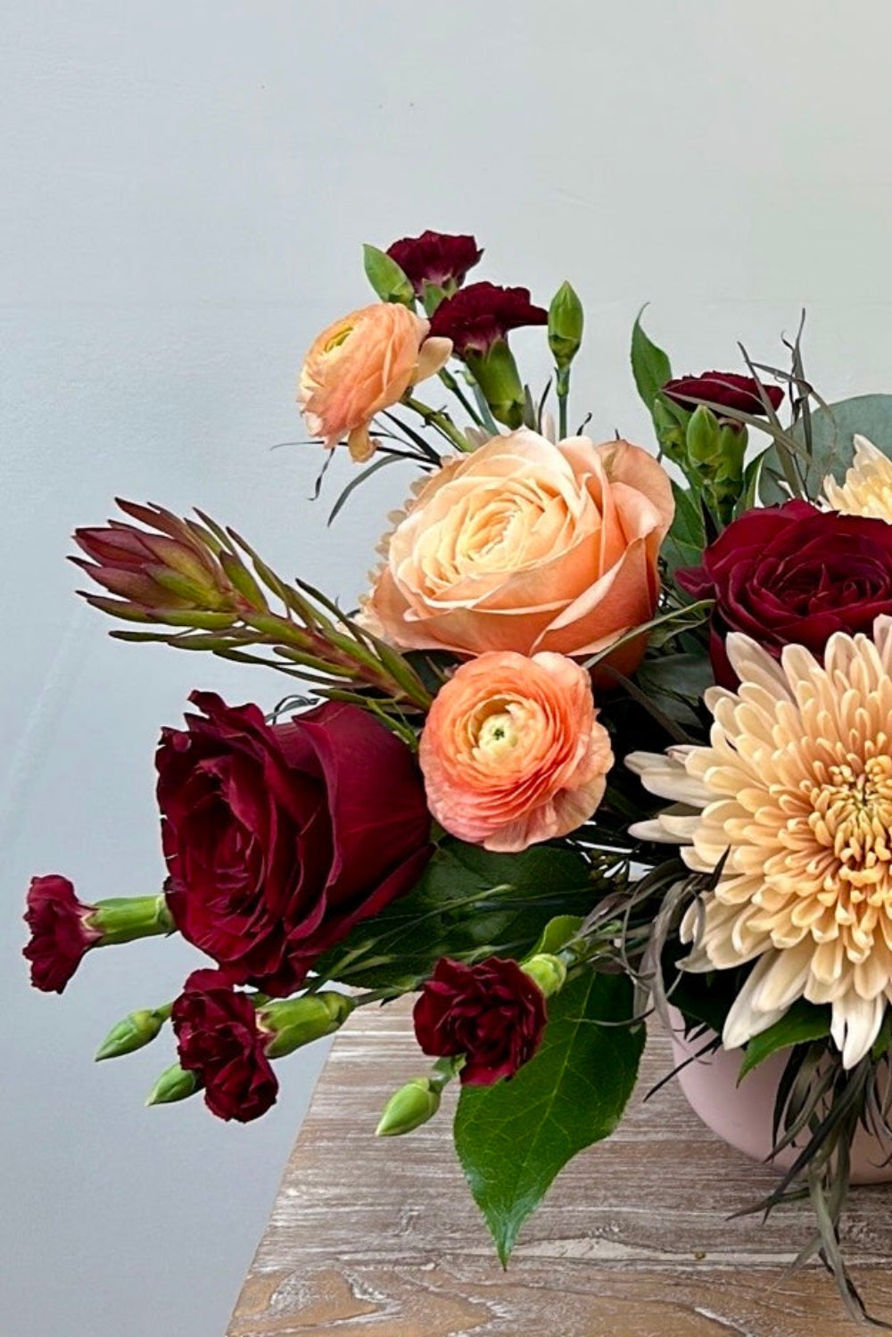 Close‑up of the Blush &amp; Bordeaux Bloom Vase showing burgundy roses, café latte cremones, peach ranunculus, blush roses, and burgundy foliage.
