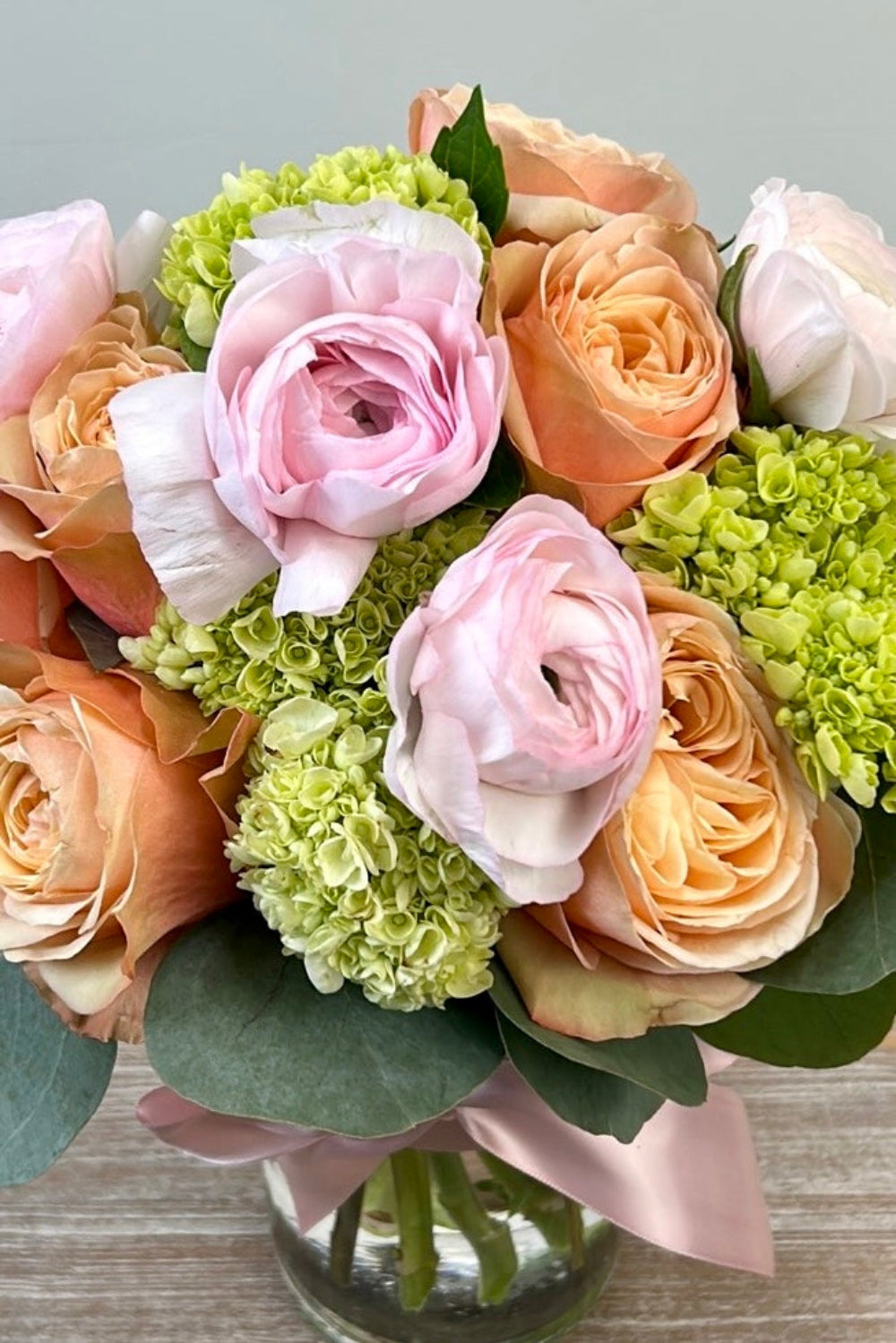 Close‑up of Pastel Petal Kiss showing pale pink ranunculus, peachy‑pink Kahala roses, soft green hydrangea, and eucalyptus details.