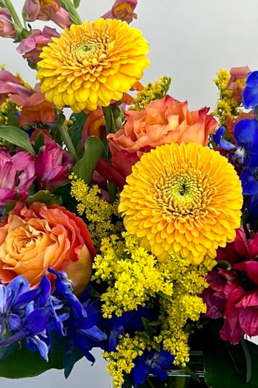 Close-up of vibrant mixed flower bouquet showing blue delphinium, orange roses, yellow germinis, hot pink alstroemeria, and apricot snapdragons