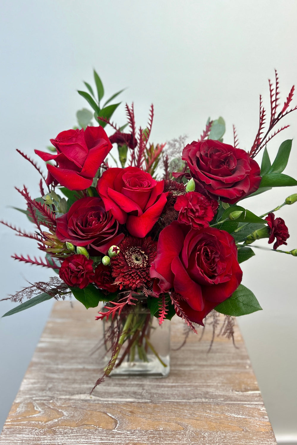 Crimson Obsession bouquet with Black Pearl roses, red Freedom roses, red cremones, burgundy pixie carnations, and red foliage arranged in a rectangular glass vase.