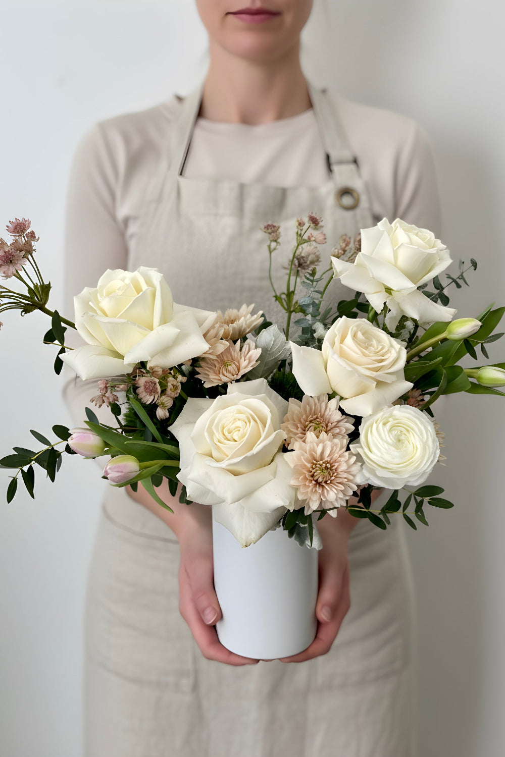 Person holding a bouquet of white and beige flowers in a white vase against a plain background, roses, tulips, ranunculus