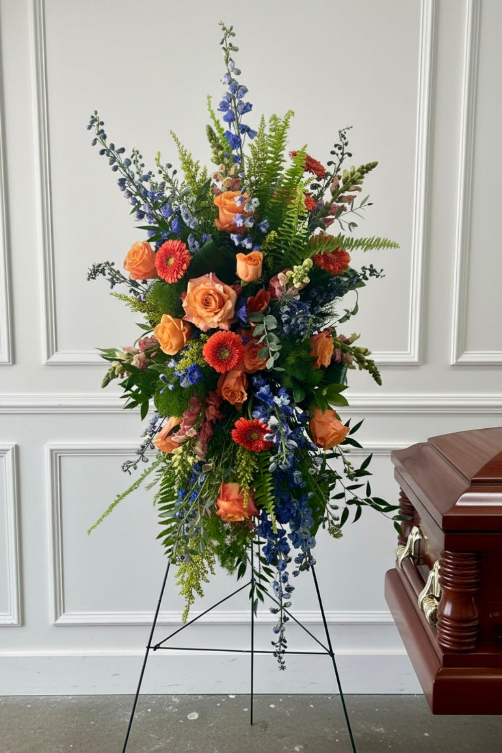 bright standing memorial funeral flower spray with roses, delphinium, and other summery flowers, suitable for a man, more masculine funeral flowers.