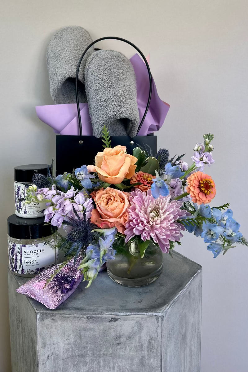 Lavender Embrace gift set with petite bouquet in light blue, lavender and blush flowers, gray fluffy slippers, lavender eye pillow, lavender foot scrub, and foot balm displayed together