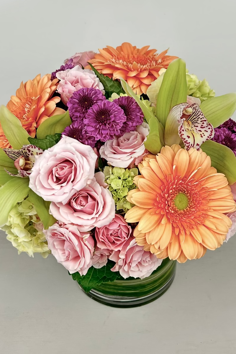 A vibrant bouquet of flowers featuring roses, spray roses, hydrangea, gerbera daisy, orchids, and ranunculus in pastel and bright colors, arranged in a leaf-wrapped vase.