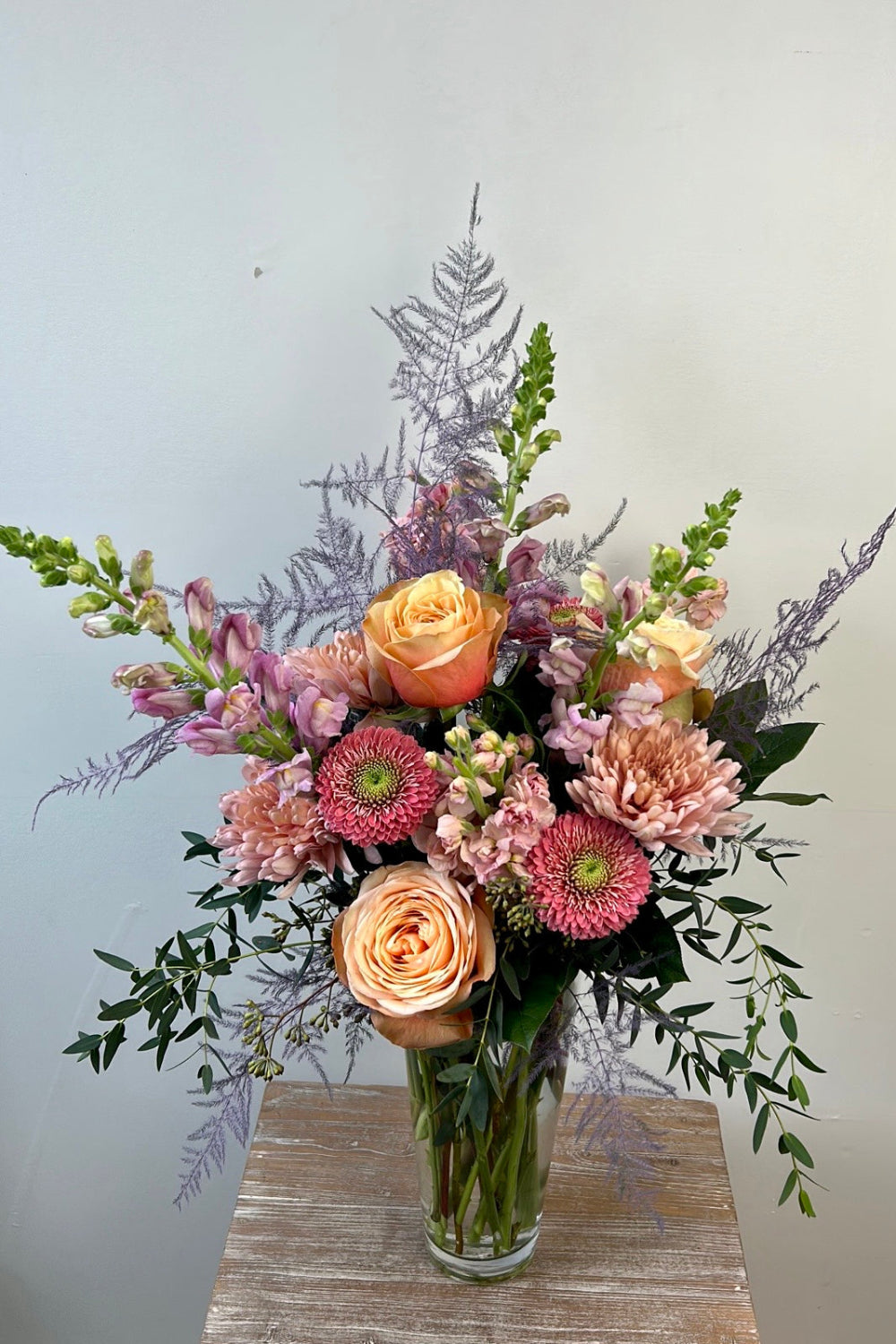 Pastel Hearts Bloom Vase with lavender snapdragons, pink roses, peach cremones, pink gerbera daisies, peach stock, and eucalyptus arranged in a tall glass vase