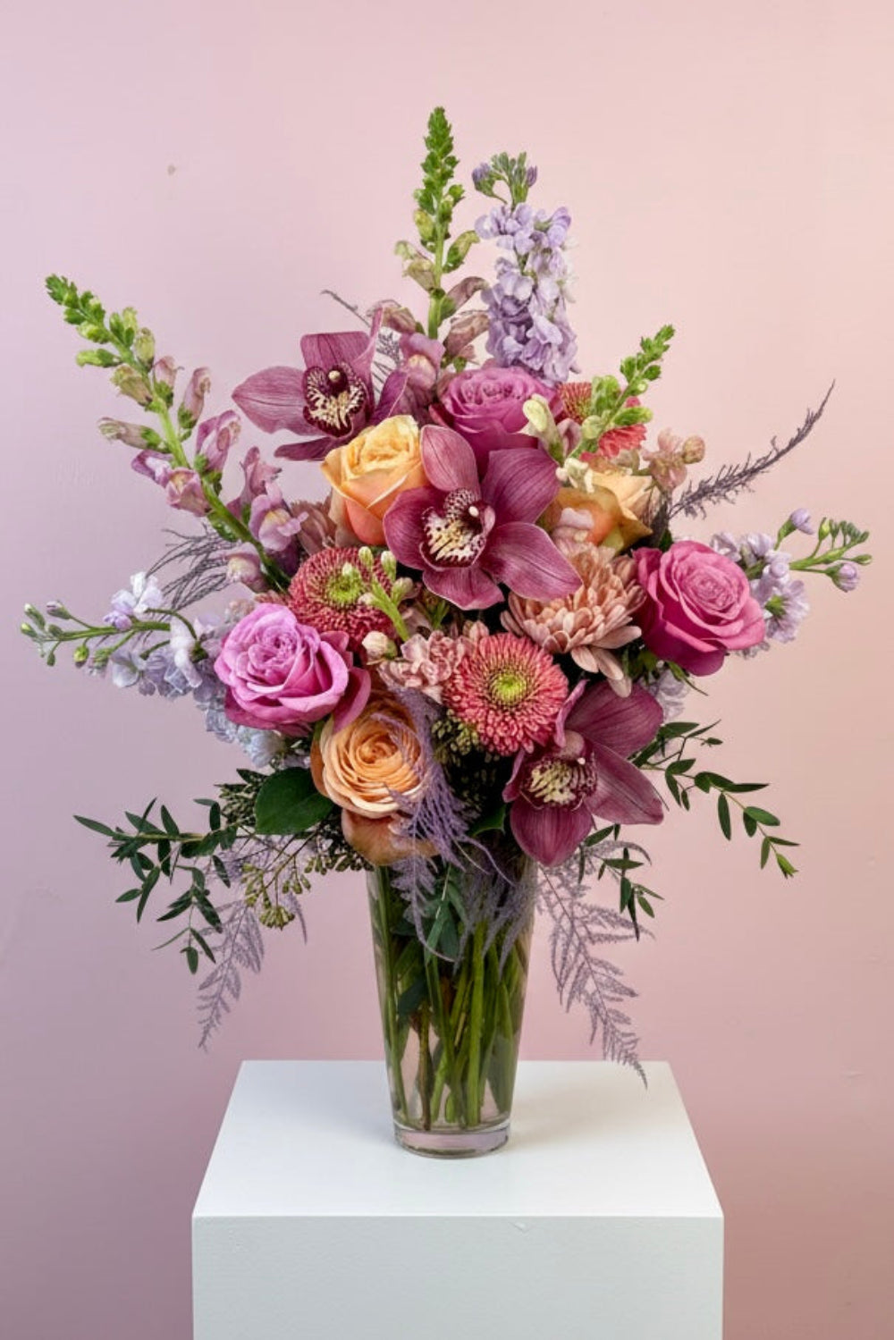 Pastel Hearts Bloom Vase with lavender snapdragons, pink roses, peach cremones, pink gerbera daisies, peach stock, and eucalyptus arranged in a tall glass vase with orchids in luxe version