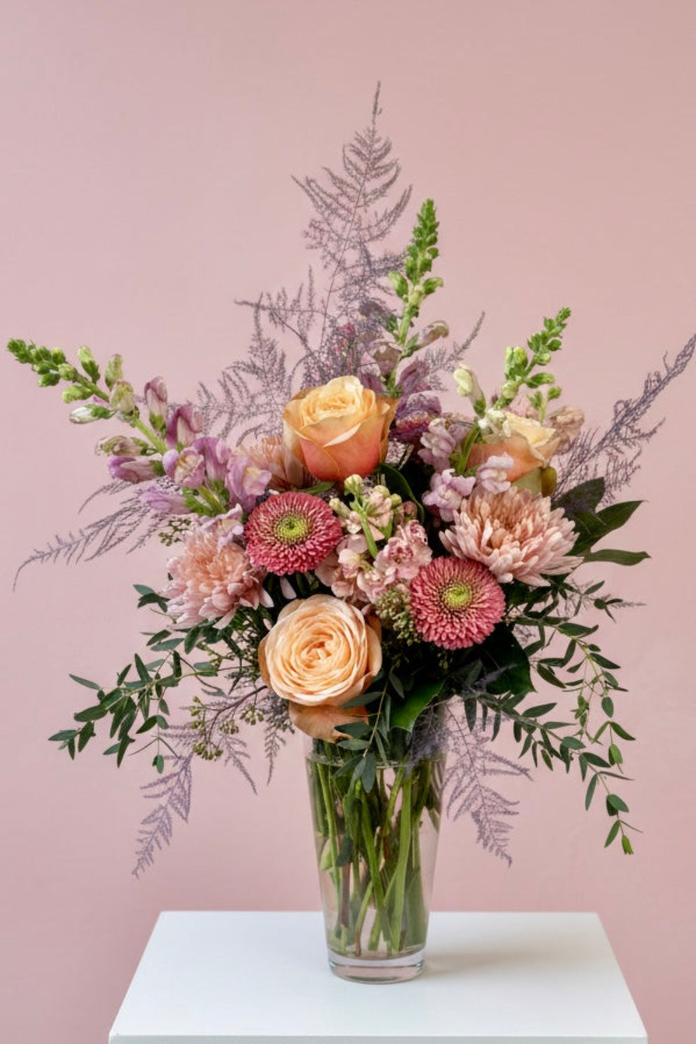 Pastel Hearts Bloom Vase with lavender snapdragons, pink roses, peach cremones, pink gerbera daisies, peach stock, and eucalyptus arranged in a tall glass vase.