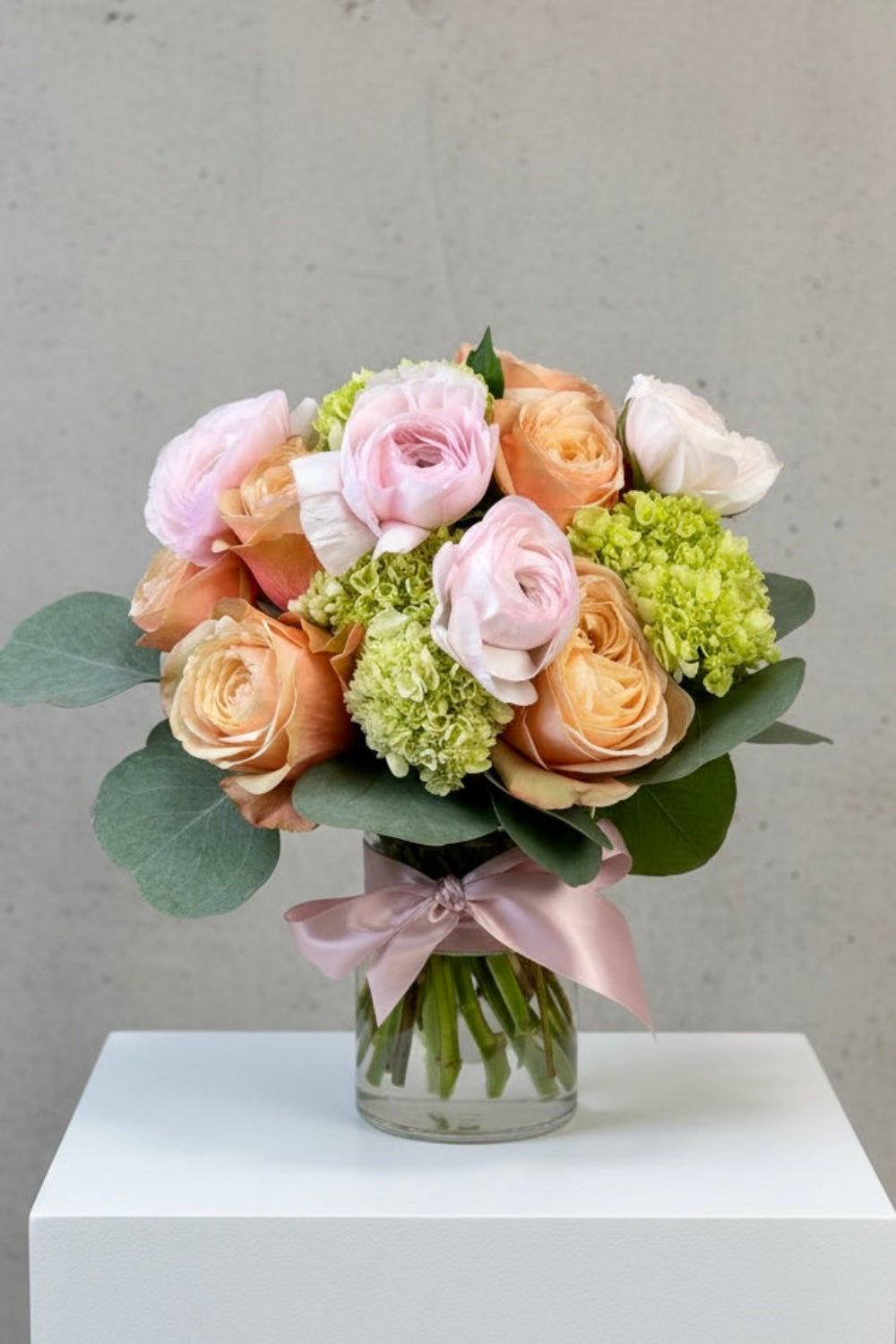 Pastel Petal Kiss bouquet with peachy‑pink Kahala roses, soft green hydrangea, pale pink ranunculus, and silver dollar eucalyptus arranged in a tight, lush style with a pale pink ribbon.