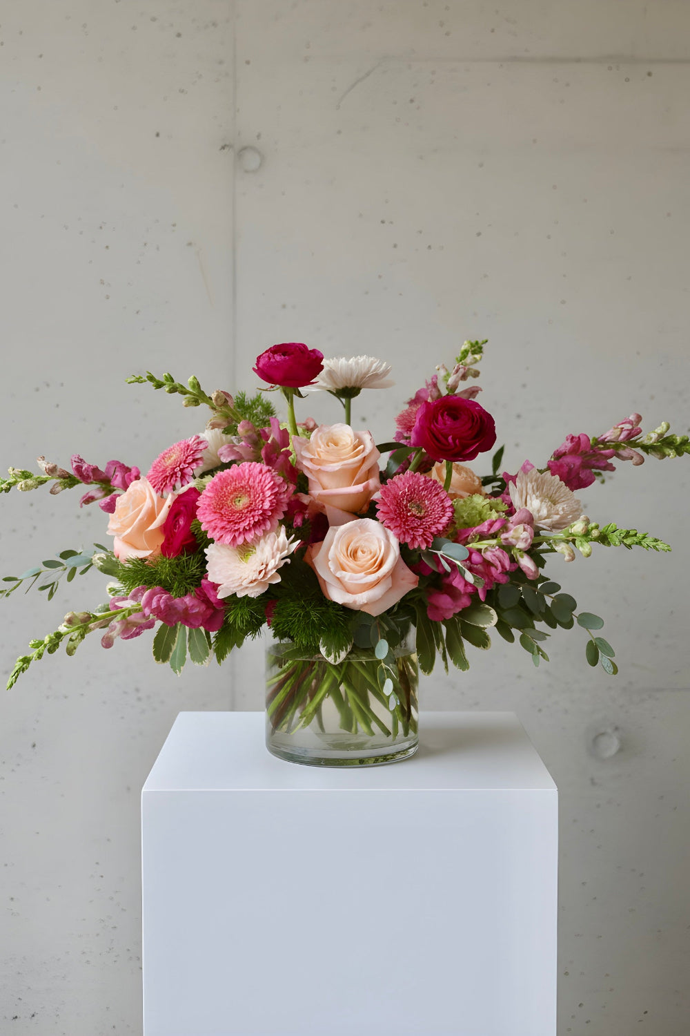 A floral arrangement in a clear glass vase sitting on a white pedestal against a light gray concrete wall. The bouquet includes blush pink roses, deep pink ranunculus, burgundy ranunculus, white daisies, and green foliage.