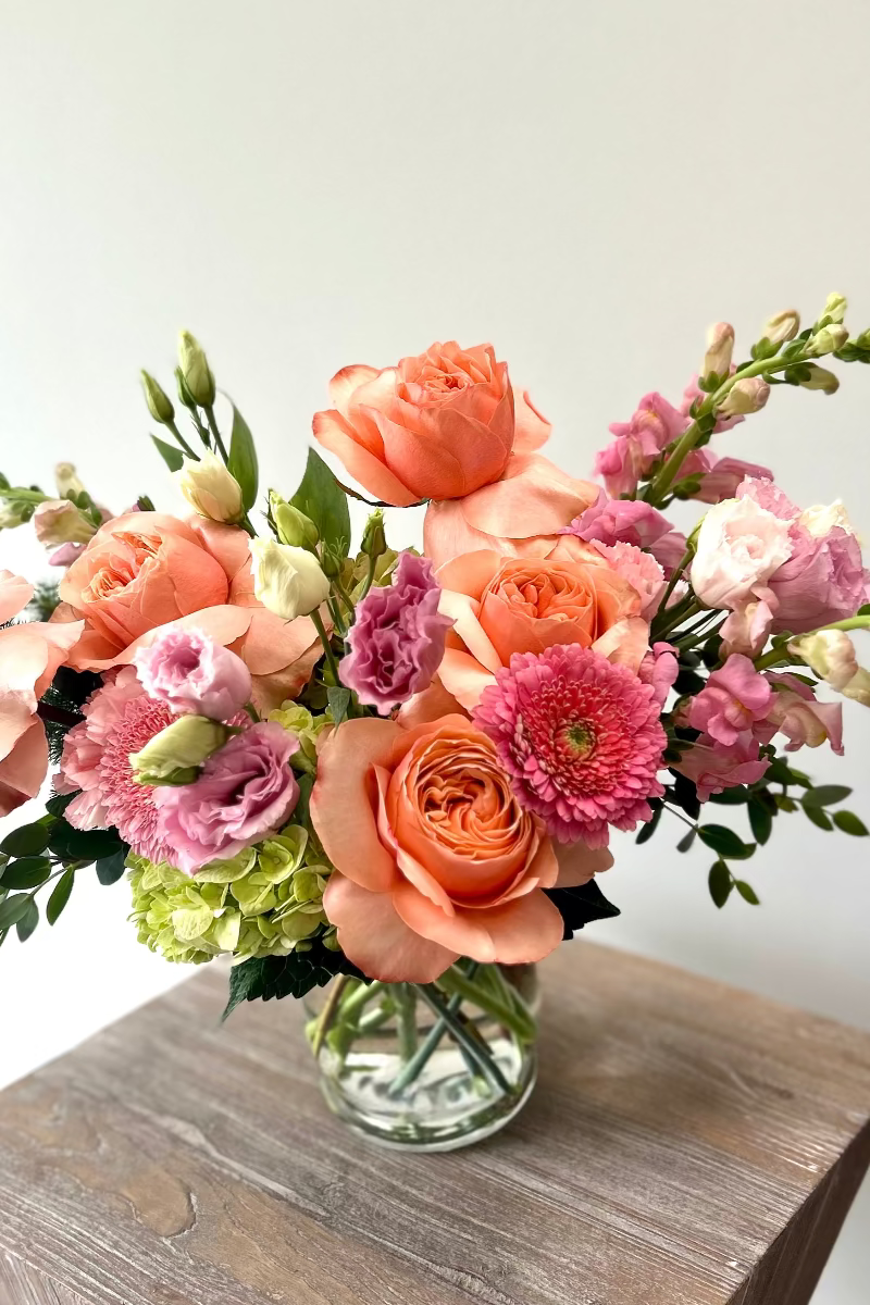 Bouquet of pink and blush flowers in a clear vase on a wooden surface with a light gray background