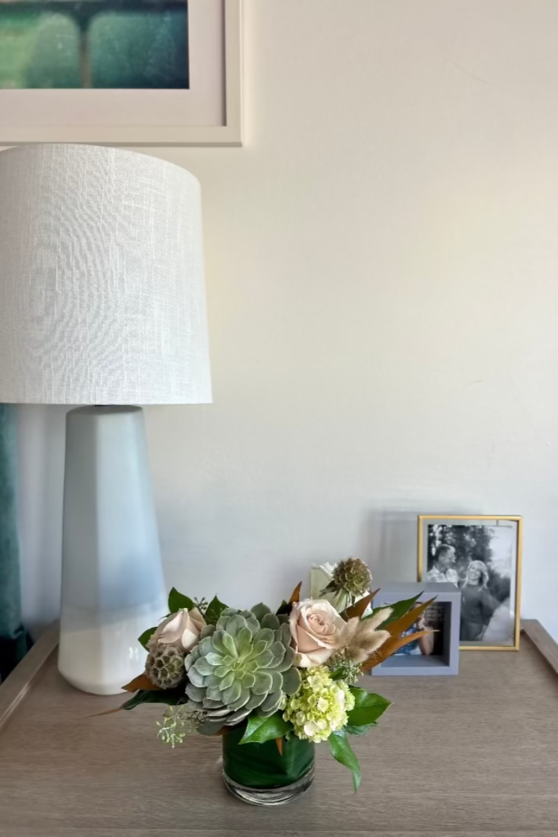Stillness and Sage petite bouquet placed on a bedside table, featuring soft green hydrangeas, tan roses, and sculptural foliage in a calming, restful setting