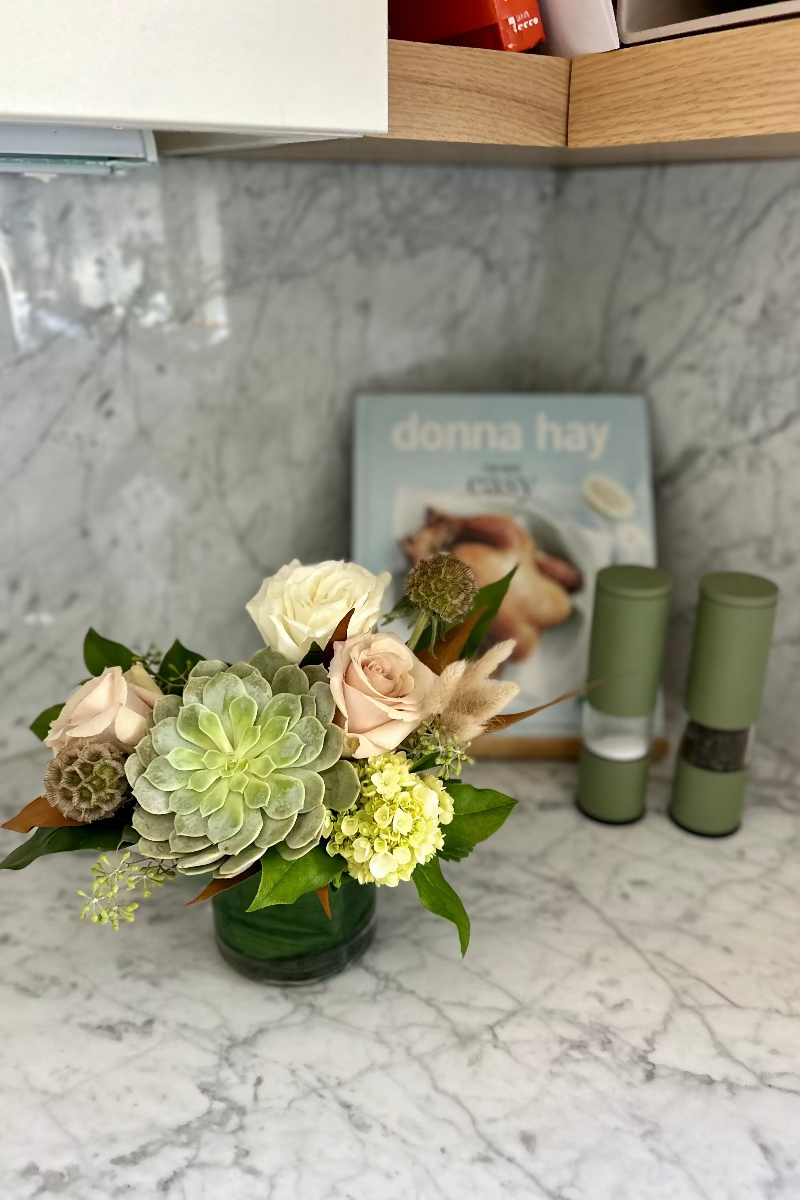 Stillness and Sage petite bouquet on a kitchen counter beside an open cookbook, showcasing neutral blooms and soft greens in a warm, everyday moment
