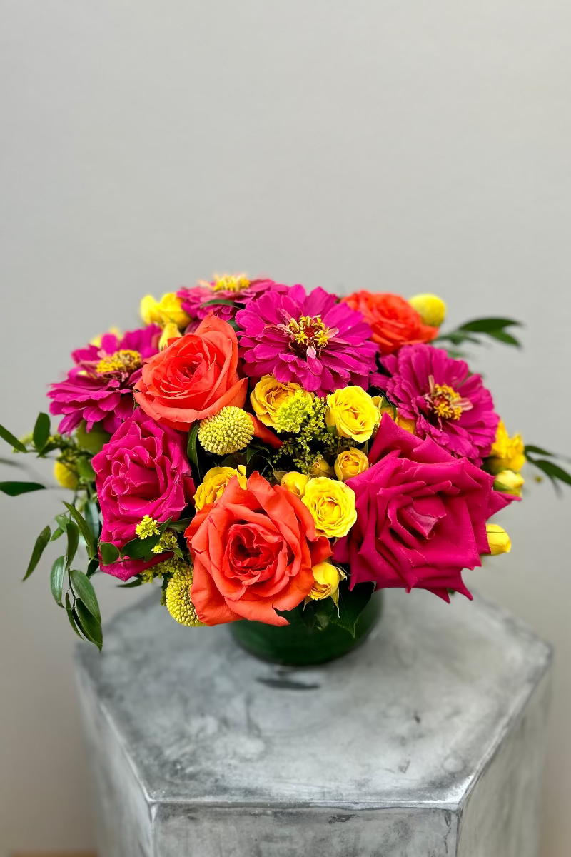Sunpop signature bouquet with bright orange and hot pink roses, hot pink gerbera daisies, yellow spray roses, and dark green foliage in a clear glass vase
