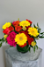 Sunpop signature bouquet with bright yellow roses, orange gerbera daisies, hot pink spray roses, and dark green foliage in a clear glass vase