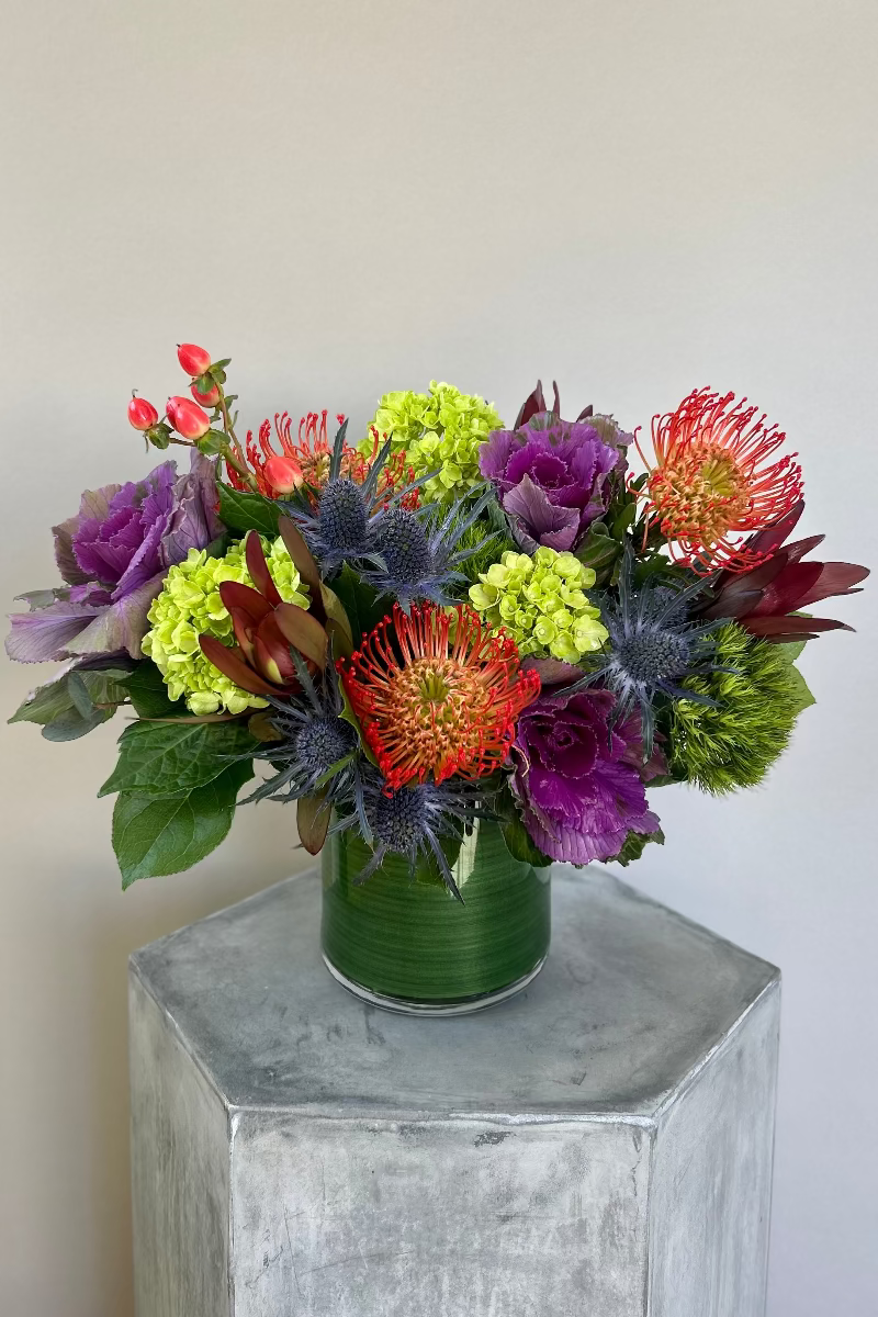 Unique bouquet with orange protea, purple ornamental kale, blue thistle, green hydrangea, and orange hypericum berries
