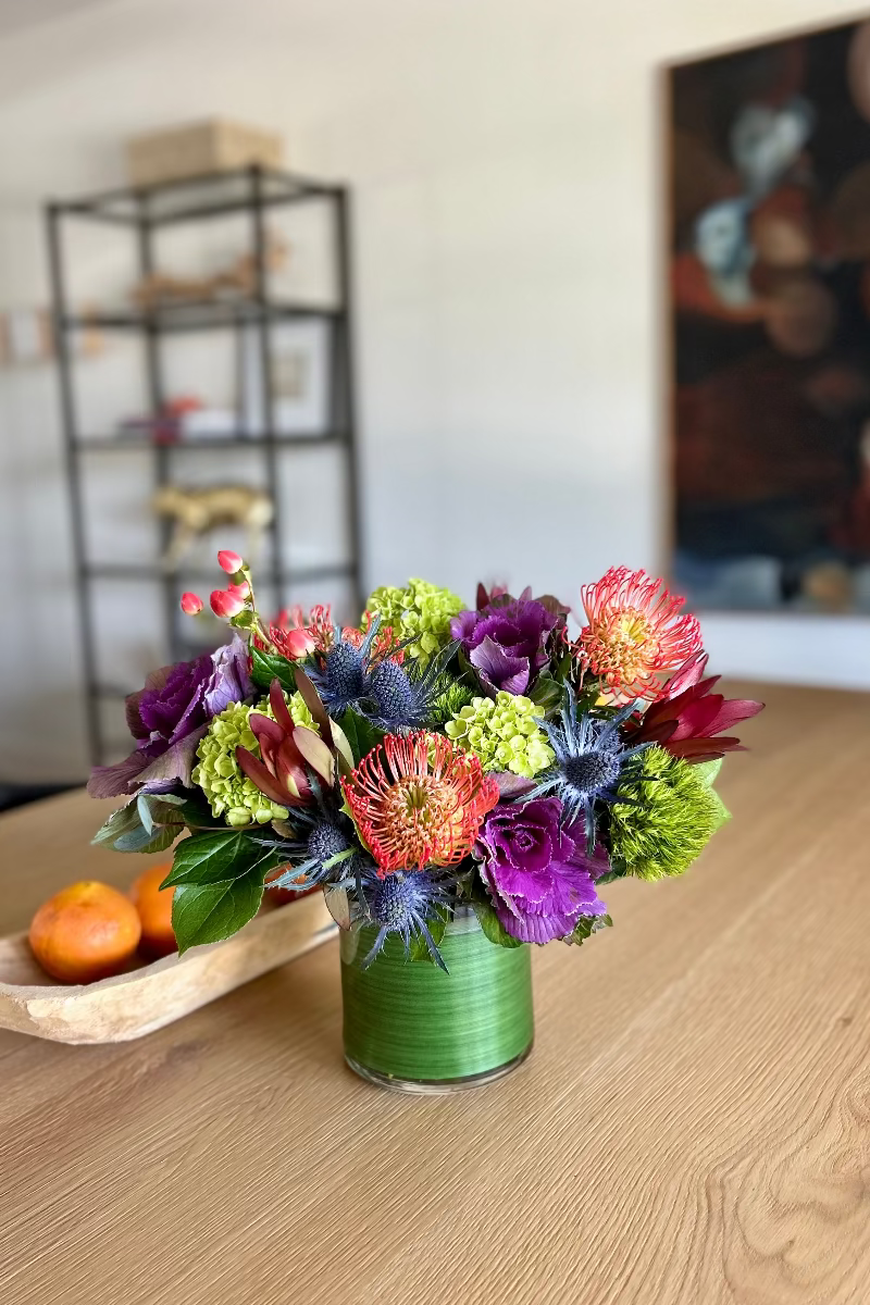 Botanic Curio bouquet displayed on a dining table, showcasing textured protea, ruffled ornamental kale, berries, thistle, and lush hydrangea in natural light