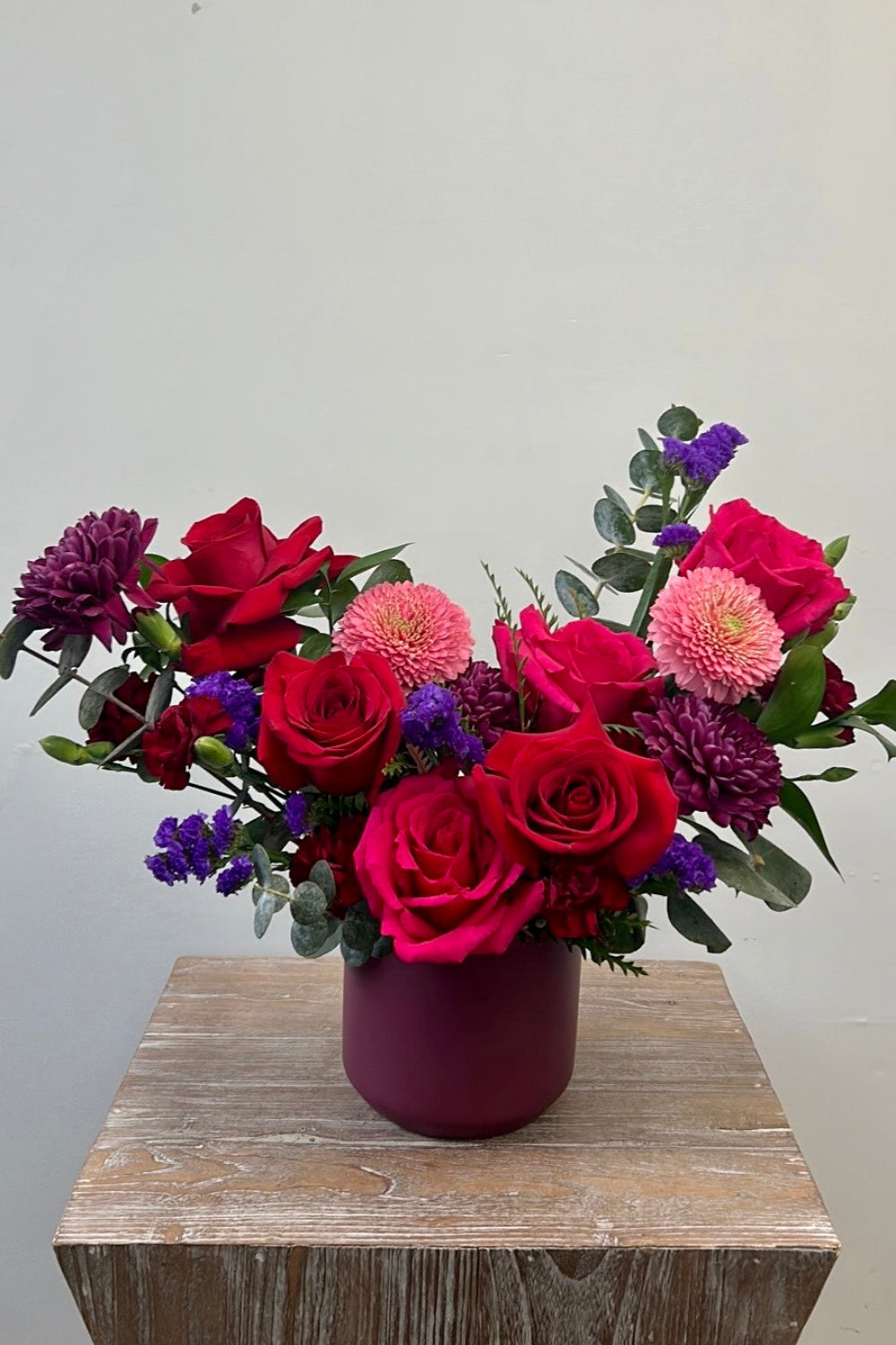 Valentine Color Crush bouquet with red roses, hot pink roses, purple cremones, pink germinis, statice, and red pixie carnations arranged in a plum ceramic vase