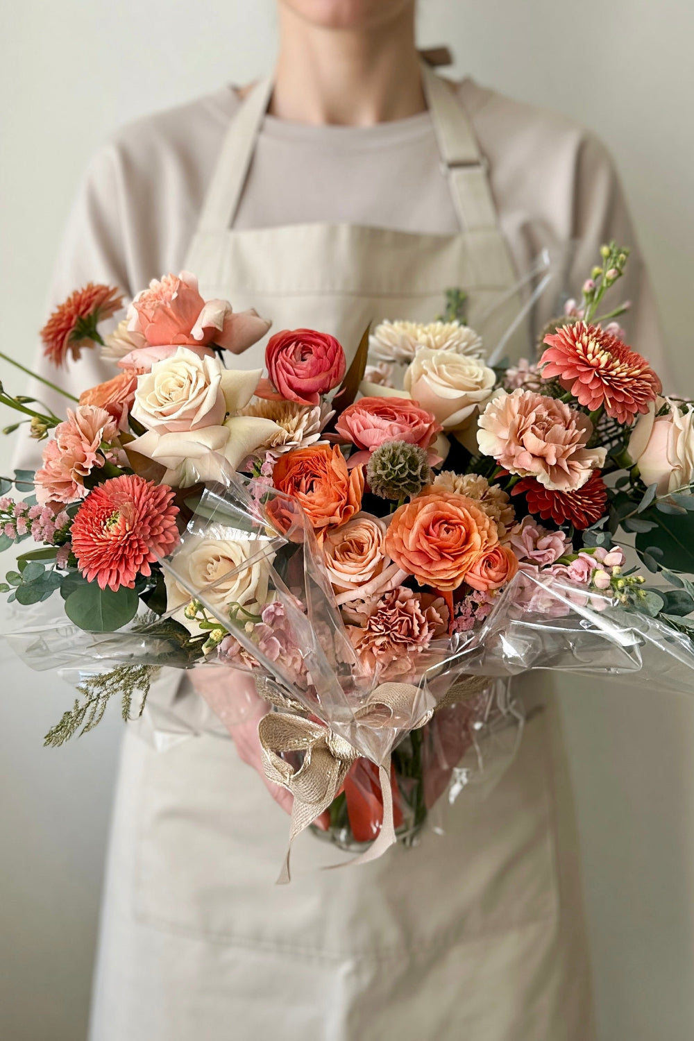  Florist holding the Premium Warm Embrace bouquet with peach, coral, and caramel flowers wrapped in cellophane and finished with a bow