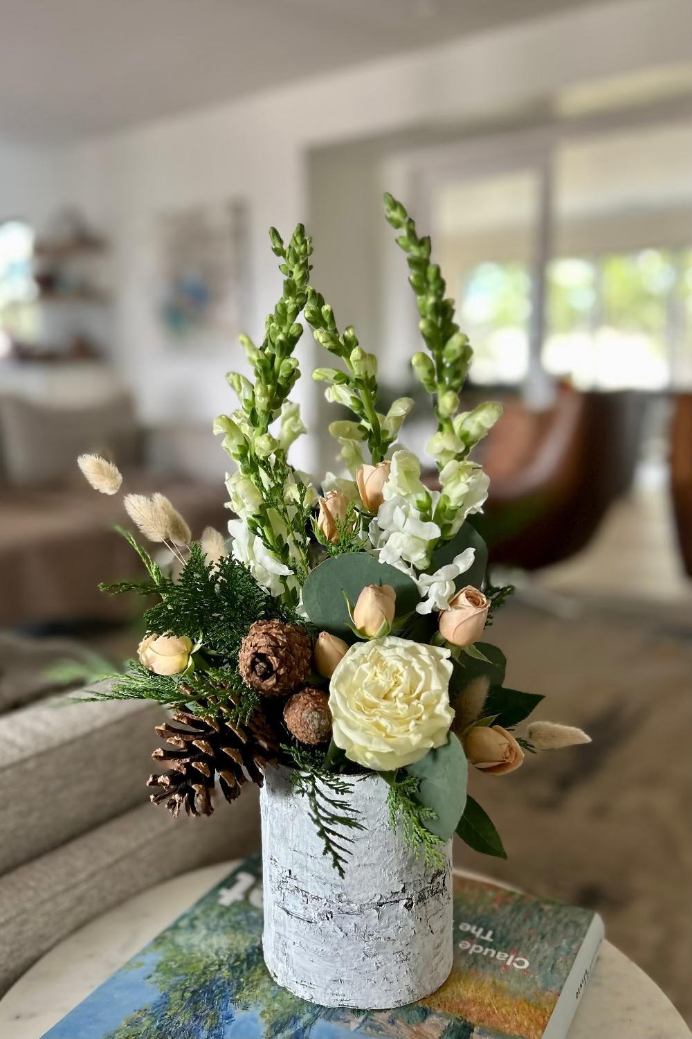 Winter Whisper Bouquet displayed in cozy living room decor, featuring birch bark vase with evergreens, roses, orchids, snapdragons, pinecones, and textured foliage