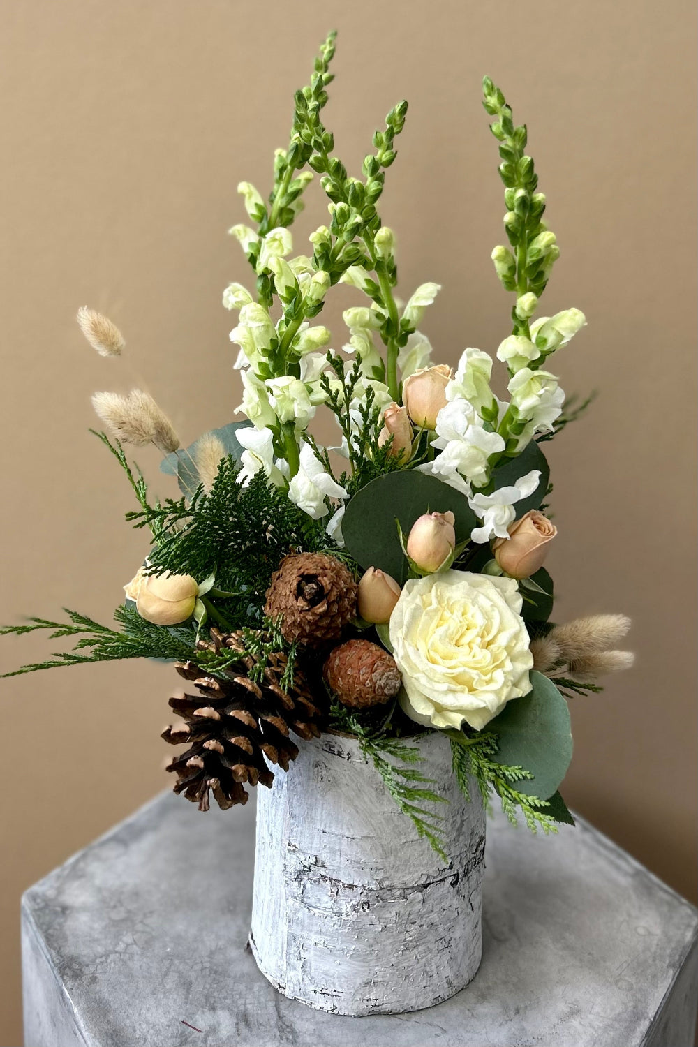 Winter Whisper Bouquet in white birch bark vase with evergreens, eucalyptus, pinecones, white snapdragons, tan spray roses, bunny tail grass, orchids, and white rose
