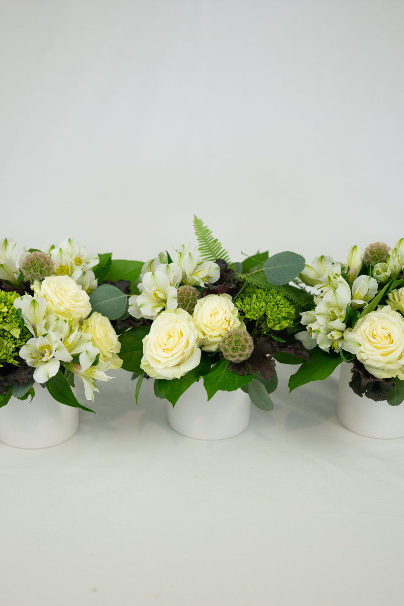 Three white rectangular containers with a modern trio table centerpiece arrangement that includes white roses, alstromeria, hydrangea, and green foliage.