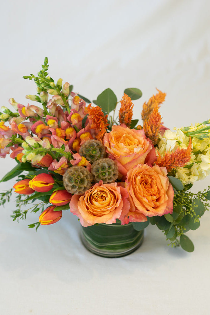 A floral arrangement featuring roses, tulips, snapdragons, and stock with seasonal foliage and botanical accents in shades of orange, yellow, and green.