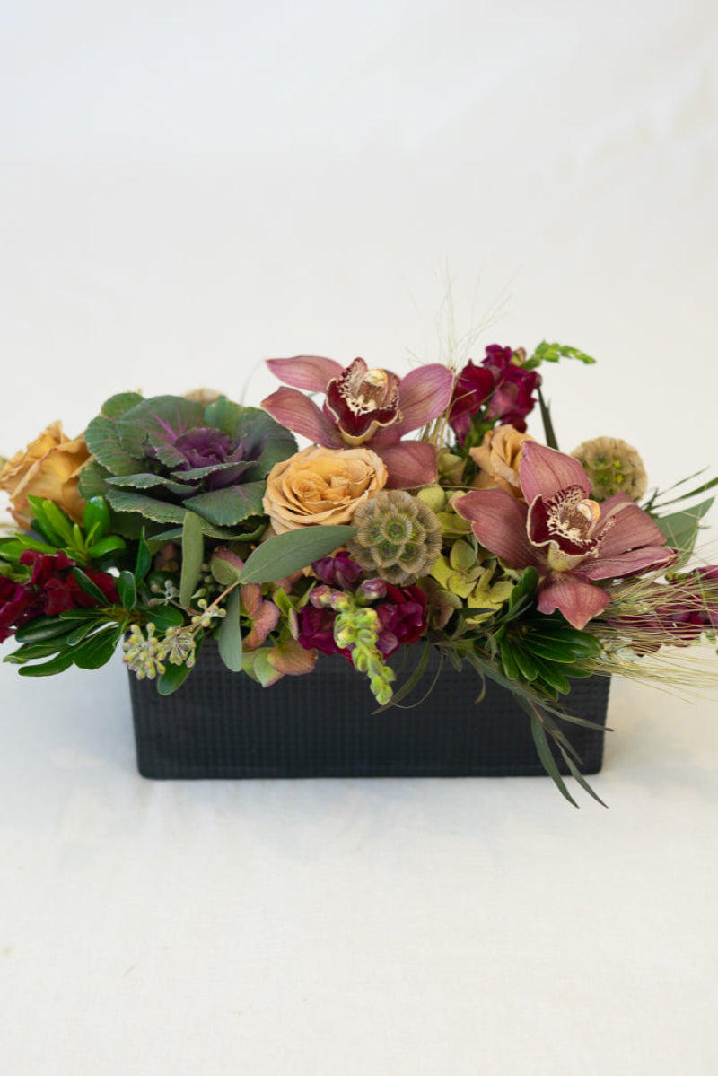 A textural table centerpiece featuring a variety of flowers including roses, orchids, and foliage, arranged in a matte black box.
