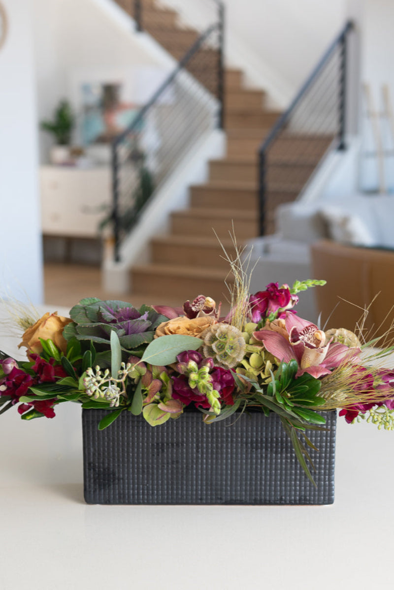 A textural table centerpiece featuring a variety of flowers including roses, orchids, and foliage, arranged in a matte black box.