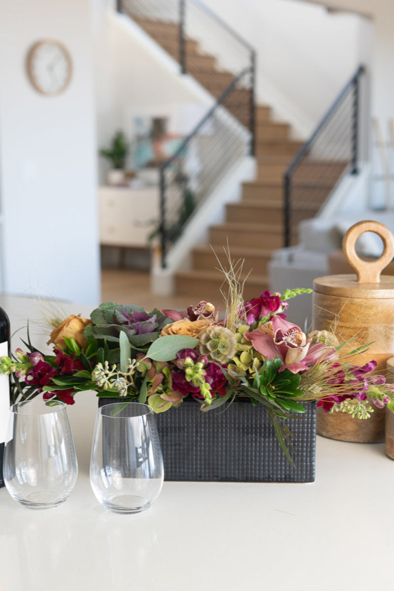 A textural table centerpiece featuring a variety of flowers including roses, orchids, and foliage, arranged in a matte black box. on island