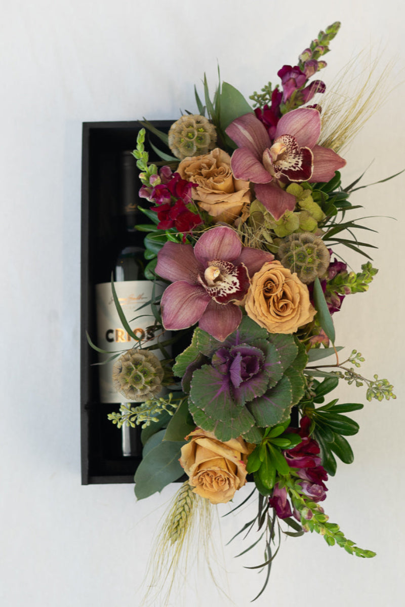 A floral arrangement featuring a variety of flowers including orchids and roses, with a keepsake box of red wine.