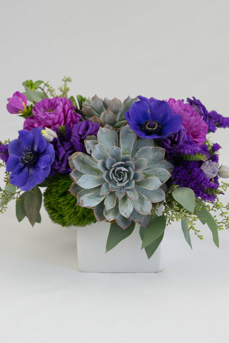 A floral arrangement featuring purple flowers and succulents in a matte white container.