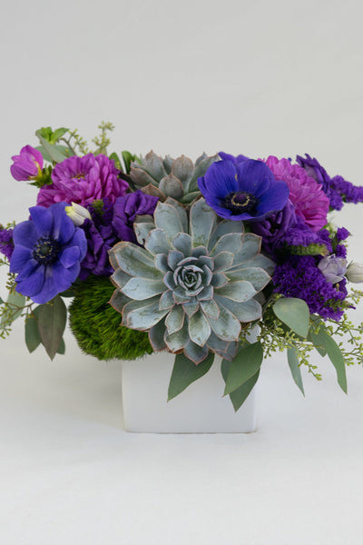 A floral arrangement featuring purple flowers and succulents in a matte white container.